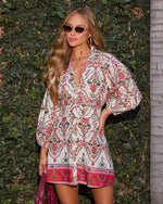 A woman wearing a bohemian-style white mini dress with red and green floral patterns, standing in front of a green hedge.