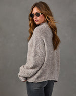 Woman in oversized taupe boucle knit sweater and gray jeans, glancing over her shoulder in gold-framed sunglasses and hoop earrings