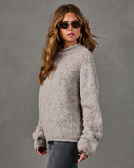 Woman wearing an oversized taupe boucle knit sweater with gold hoop earrings, oval sunglasses, and gray jeans against a gray backdrop