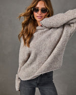 Woman wearing an oversized taupe boucle knit sweater with gold oval sunglasses, hoop earrings and dark gray jeans