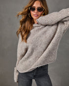 Woman wearing an oversized taupe boucle knit sweater with gold oval sunglasses, hoop earrings and dark gray jeans