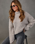 Woman in oversized gray boucle turtleneck sweater, dark skinny jeans and gold sunglasses, seated on a black stool