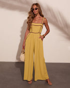 Woman wearing mustard yellow wide-leg jumpsuit with burgundy trim, straw bag, cork heels, and gold accessories
