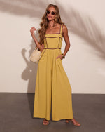 Woman wearing mustard yellow wide-leg jumpsuit with burgundy piping, straw bag, gold bangles, and nude sandals