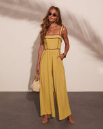 Woman wearing chartreuse wide-leg jumpsuit with brown piping, straw bag, strappy sandals, and oval sunglasses