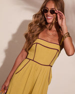 Woman in mustard yellow sundress with burgundy trim, smiling, wearing oval sunglasses and gold bangles, summer fashion