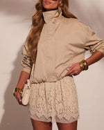 Woman wearing beige windbreaker jacket with cream lace mini skirt, gold bangles, and white clutch bag