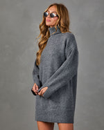 A woman wearing an oversized gray turtleneck sweater dress, styled with round sunglasses, standing against a textured gray studio background.