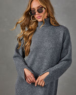 A woman wearing a heathered gray knit turtleneck sweater dress, styled with gold hoop earrings and sunglasses, against a textured gray studio background.
