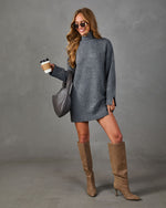A woman models a gray knit turtleneck sweater dress, styled with brown knee-high suede boots, a gray quilted shoulder bag, and sunglasses while holding a coffee cup against a gray studio backdrop.