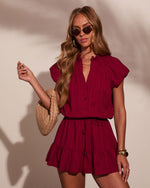Woman wearing burgundy ruffle romper with button detail and drawstring waist, styled with woven bag and tinted sunglasses