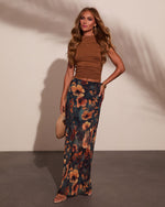 Woman wearing brown ruched crop top and teal floral maxi skirt with gold accessories and woven clutch bag