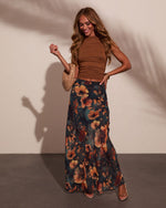 Woman wearing brown ruched crop top and dark floral maxi skirt with amber flowers, holding woven clutch, nude heels