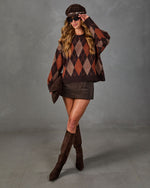 A woman models a brown argyle sweater, matching brown skirt, brown over-the-knee boots, and a brown cap against a textured gray studio backdrop.