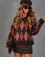 A woman wearing a brown and rust argyle knit sweater, a matching brown hat, sunglasses, and a brown mini skirt, posing against a textured gray studio background.