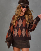 A woman wearing a brown and rust argyle knit sweater, a matching brown hat, sunglasses, and a brown mini skirt, posing against a textured gray studio background.