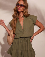 Woman wearing olive green ruffle mini dress with drawstring waist, aviator sunglasses, woven bag, and gold jewelry