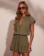 Woman wearing olive green cotton gauze romper with ruffle details, drawstring waist, and cap sleeves, styled with aviator sunglasses and gold jewelry