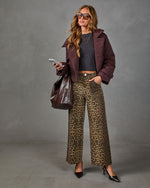 A woman models a stylish outfit featuring wide-leg leopard print trousers, a dark gray top, a brown jacket, and a large brown leather tote bag against a textured gray studio background.