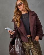 A woman wearing a quilted plum-burgundy jacket, leopard-print pants, a black top, and sunglasses, holding a phone and a large dark shoulder bag against a gray studio background.