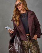 A woman wearing a quilted plum-burgundy jacket, leopard-print pants, a black top, and sunglasses, holding a phone and a large dark shoulder bag against a gray studio background.