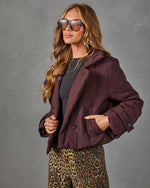 A woman wearing a dark plum-burgundy quilted jacket, leopard-print pants, a dark gray top, and sunglasses, posing against a textured gray studio background.