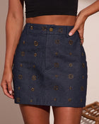 Dark navy denim mini skirt with gold floral stud embellishments, high waist, brass button closure, paired with black crop top