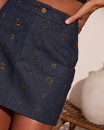 Dark navy denim mini skirt with gold sunburst stud embellishments and front patch pocket, modeled on woman