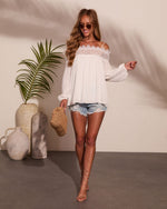 Woman wearing white lace cold-shoulder top, denim cutoff shorts, nude strappy heels, holding woven bag, boho summer style