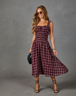 A woman modeling a sleeveless smocked midi dress with a burgundy and white plaid pattern, styled with sunglasses, a black clutch, and strappy high-heeled sandals against a textured gray studio backdrop.