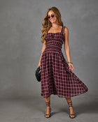 A woman modeling a sleeveless smocked midi dress with a burgundy and white plaid pattern, styled with sunglasses, a black clutch, and strappy high-heeled sandals against a textured gray studio backdrop.