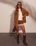 Woman in western chic outfit: tan chore coat, white tee, brown lace-trim shorts, dark cowboy boots, holding brown bag