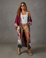 Woman wearing a long burgundy mountain-print cardigan with white tank, tan leather shorts, suede knee-high boots and woven clutch