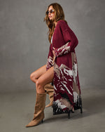 Woman wearing a long burgundy mountain-print cardigan with tan suede knee-high boots and round sunglasses, seated on a stool
