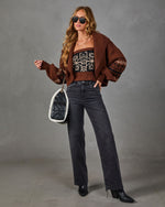 A woman models a brown knit cardigan with a geometric pattern, worn over a patterned top and paired with dark gray jeans and black boots, holding a black quilted handbag against a gray studio background.
