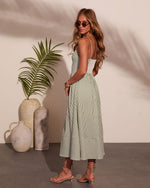 Woman in green striped midi dress with smocked back, spaghetti straps, lace-up heels, and sunglasses, posed by tropical decor