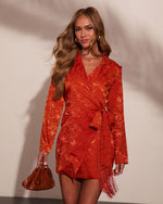 Woman wearing rust orange jacquard wrap mini dress with fringe hem and tie waist, styled with tan leather bag and gold jewelry