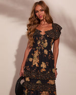 Woman wearing black floral maxi dress with ruffle details and lace-up bodice, holding woven leather clutch bag