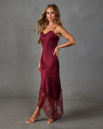 A woman models a burgundy satin slip dress with matching lace panels and sheer lace hem, styled with clear high-heeled sandals against a textured gray studio background.