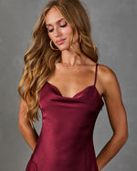 A woman models a burgundy satin slip dress with matching lace panels and sheer lace hem, styled with clear high-heeled sandals against a textured gray studio background.