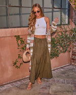 Woman in olive green tiered maxi skirt, white crop top, striped cardigan, tan sandals, holding woven bag outdoors