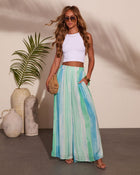 Woman wearing white crop top and blue-green striped maxi skirt with woven clutch, strappy sandals, and gold accessories