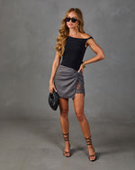 Woman wearing black off-shoulder bodysuit, gray lace-trim wrap mini skirt, strappy heels, holding black clutch, gold accessories