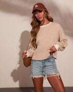 Woman wearing cream striped knit top, denim cutoff shorts, brown LA cap, gold hoops, and tan crossbody bag