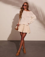 Woman wearing cream knit off-shoulder sweater, tiered mini skirt, tan heeled mules, hoop earrings, and crossbody bag