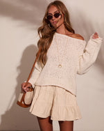 Woman wearing cream off-shoulder knit sweater, tiered mini skirt, gold accessories, and tan bucket bag, boho style outfit