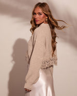 Woman wearing beige ribbed knit sweater with lace hem trim over satin slip skirt, long wavy blonde hair, looking over shoulder