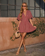 Woman in red plaid mini dress with flutter sleeves, nude strappy heels, and beige handbag posing outdoors