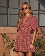 Woman in red plaid mini shirtdress with sunglasses, gold jewelry, and woven bag, posed outdoors against terracotta wall