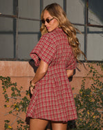 Woman wearing red plaid mini shirtdress with short sleeves, gold bangles, and sunglasses, posed outdoors against terracotta wall